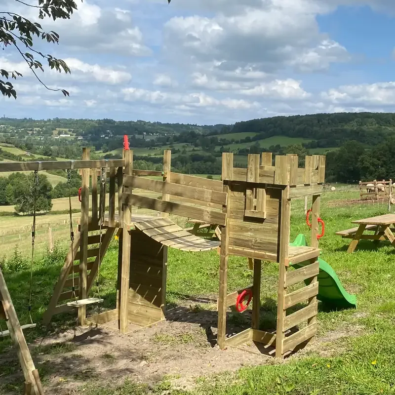 L'aire de jeux en bois : sa tour crénelée, son toboggan et sa balançoire, face aux collines du Pays d'Auge