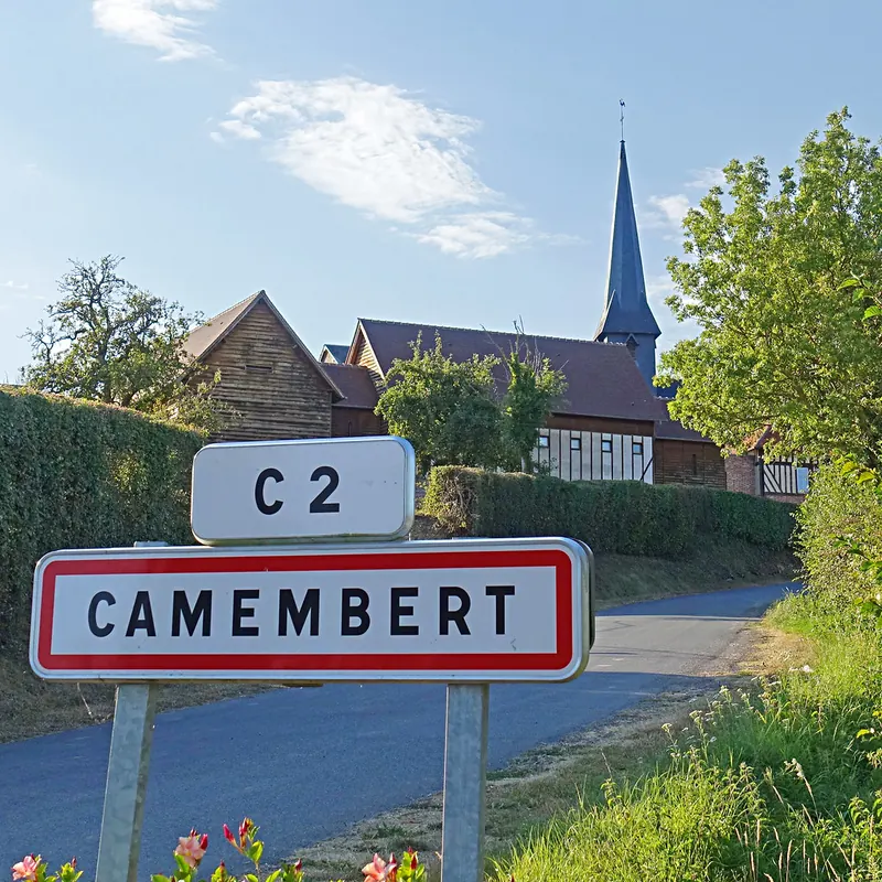 Le panneau d'entrée du village de Camembert, l'église et ses maisons à colombages derrière la haie